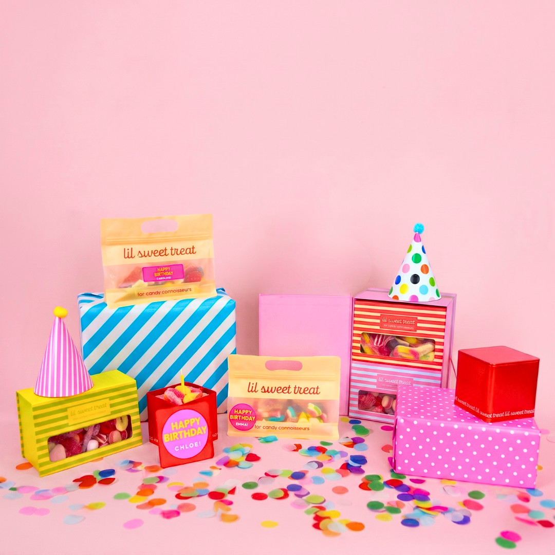 A styled arrangement of colorful gift boxes and candy packages on a pink background with confetti scattered in front. Visible items include a blue-and-white striped box, a pink box, a yellow crate, a red tin, and bags labeled 'lil sweet treat,' with party hats on some birthday presents.